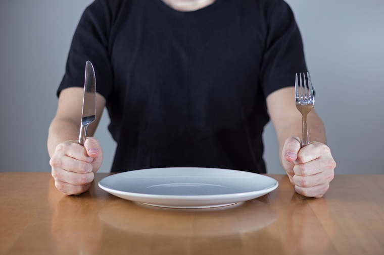 A person sitting at a dining table holding a knife and fork with a plate in between