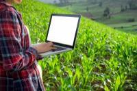 A person standing on an open agricultural field, holding a laptop and taking notes.